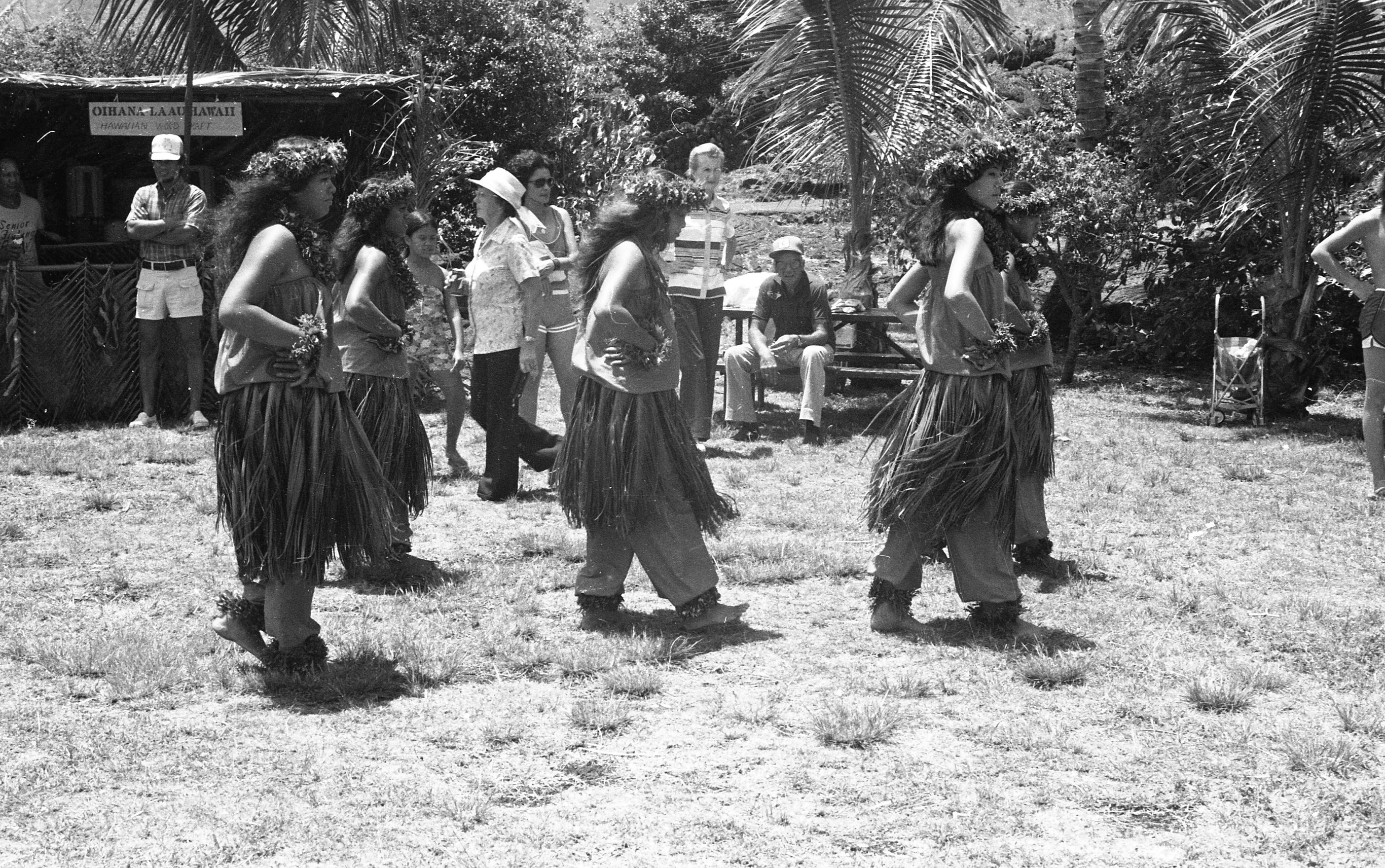 A black and white image of five women performing at an outdoor event. The five women are located in the center of the image. They are all dressed similarly. They are wearing a flower crown, lei, tube top, flower bracelets on each of their wrists, grass skirt, and flower anklets on both of their ankles. They have their hands on their hips walking towards the right side of the image. Behind the dancers there are people watching them. In the background there is a vending booth on the left side of the image. The booth is constructed out of palm leaves. There is a sign hanging down from its roof that reads, "Ohana La Au Hawaii, Hawaii Wood Craft."
