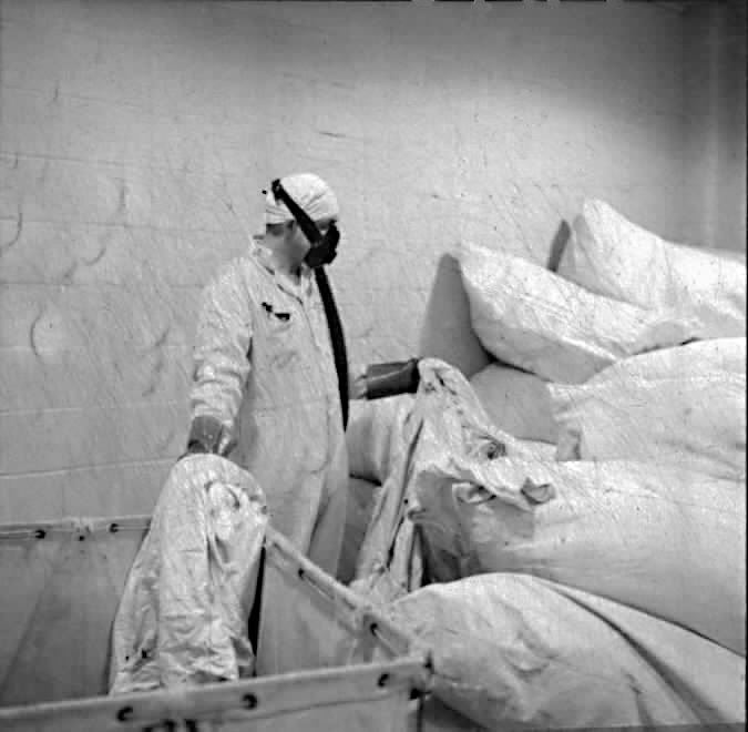 Black and white photo of a person in white coveralls and cap handling large cloth bags. 