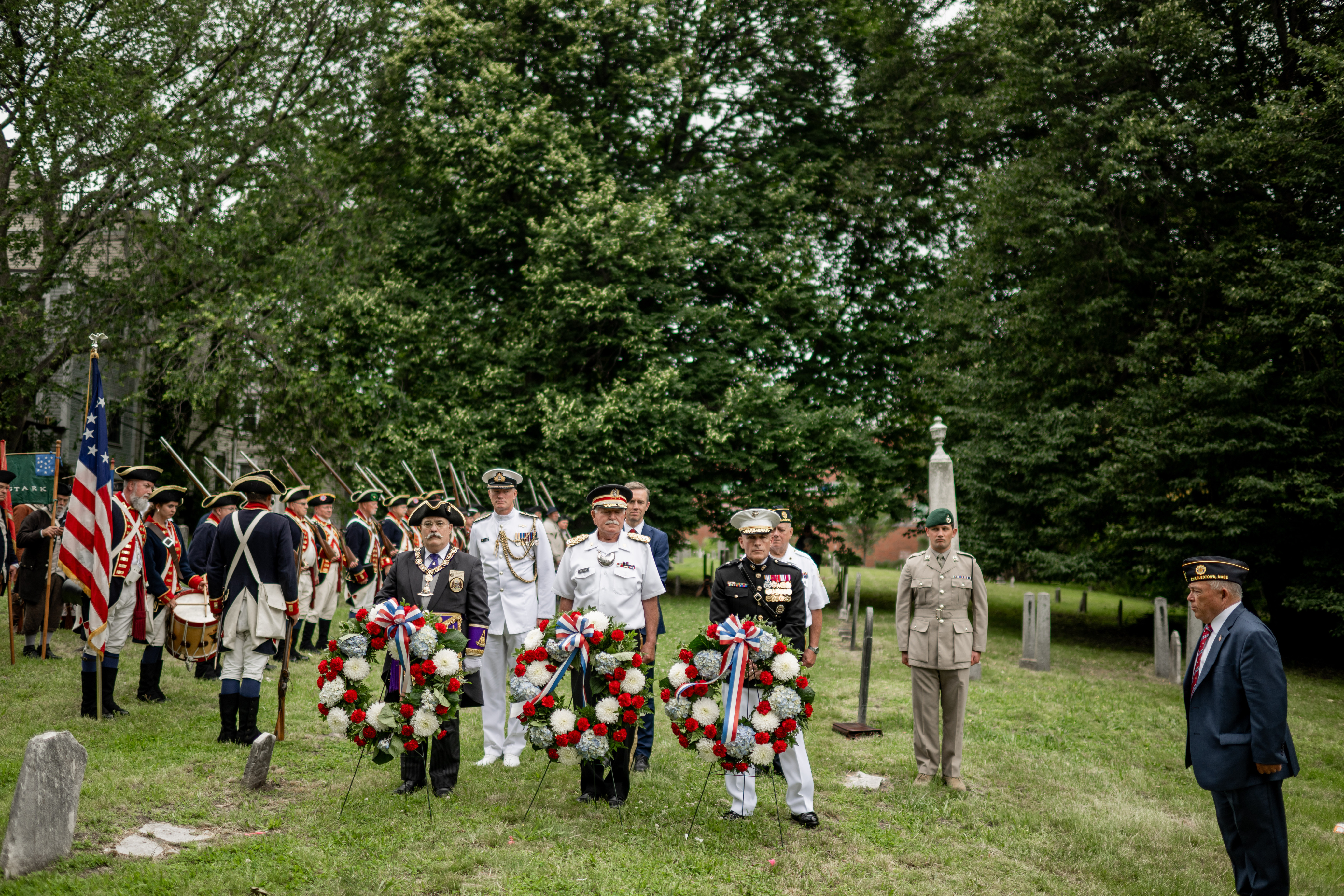 Three wreaths are on stands in a cemetery. Behind each wreath stands an individual in uniform. A militia group stands behind them off to the left. 