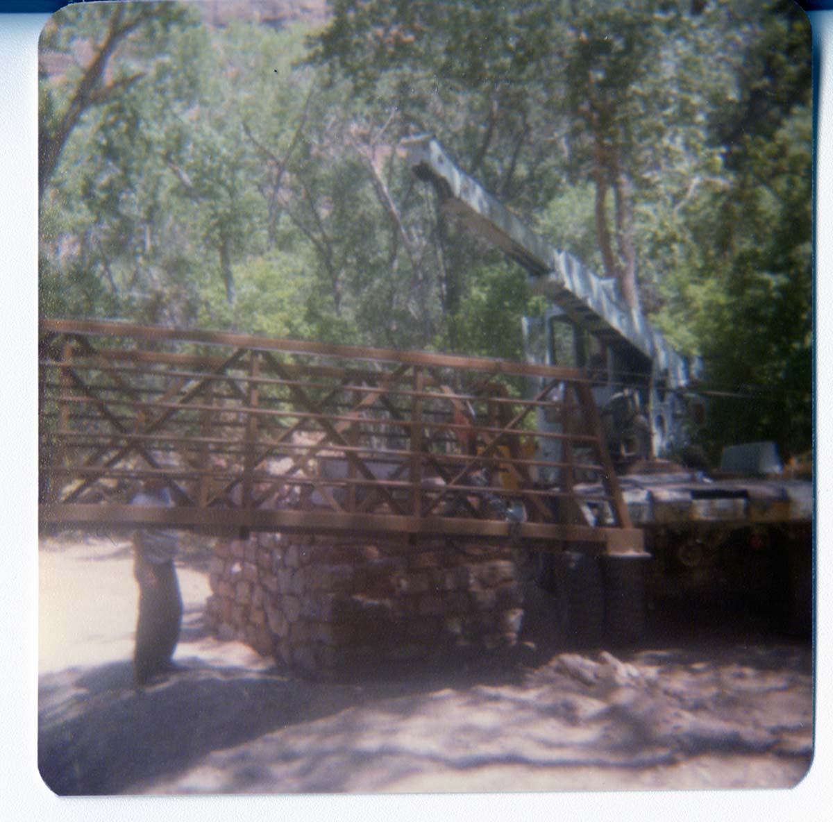 Man stands by while crane lifts one end of new Grotto footbridge onto stone abutment.