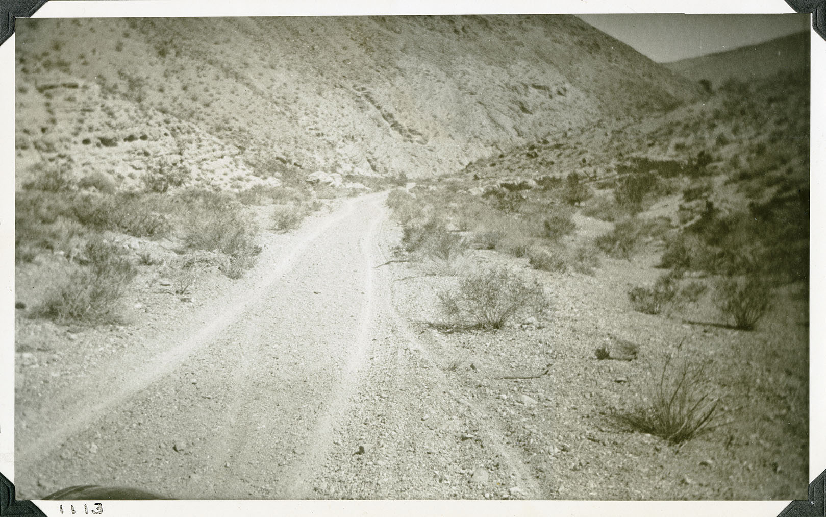 This is an historic black and white photograph from the Scotty's Castle Historic Photograph Collection, Death Valley National Park of desert canyon and wash. Two track dirt road lead down from wash. Narrow wash thick with gravel. On left a steep canyon wall. On right a hillside. Sparce vegetation leading up from road to hillsides. Number in black ink in lower left corner.