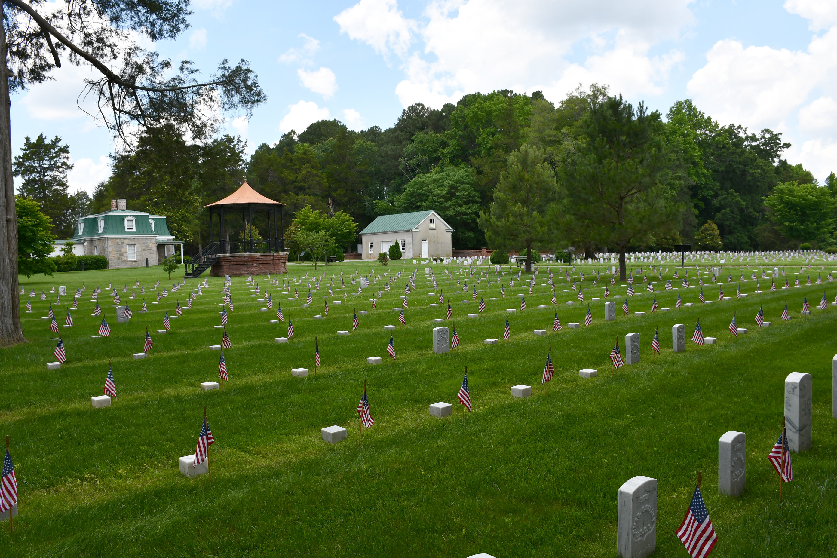 The Superintendent’s Lodge, maintenance building, and rostrum in the background and in the fore ground American flags stand in front of white headstones. 