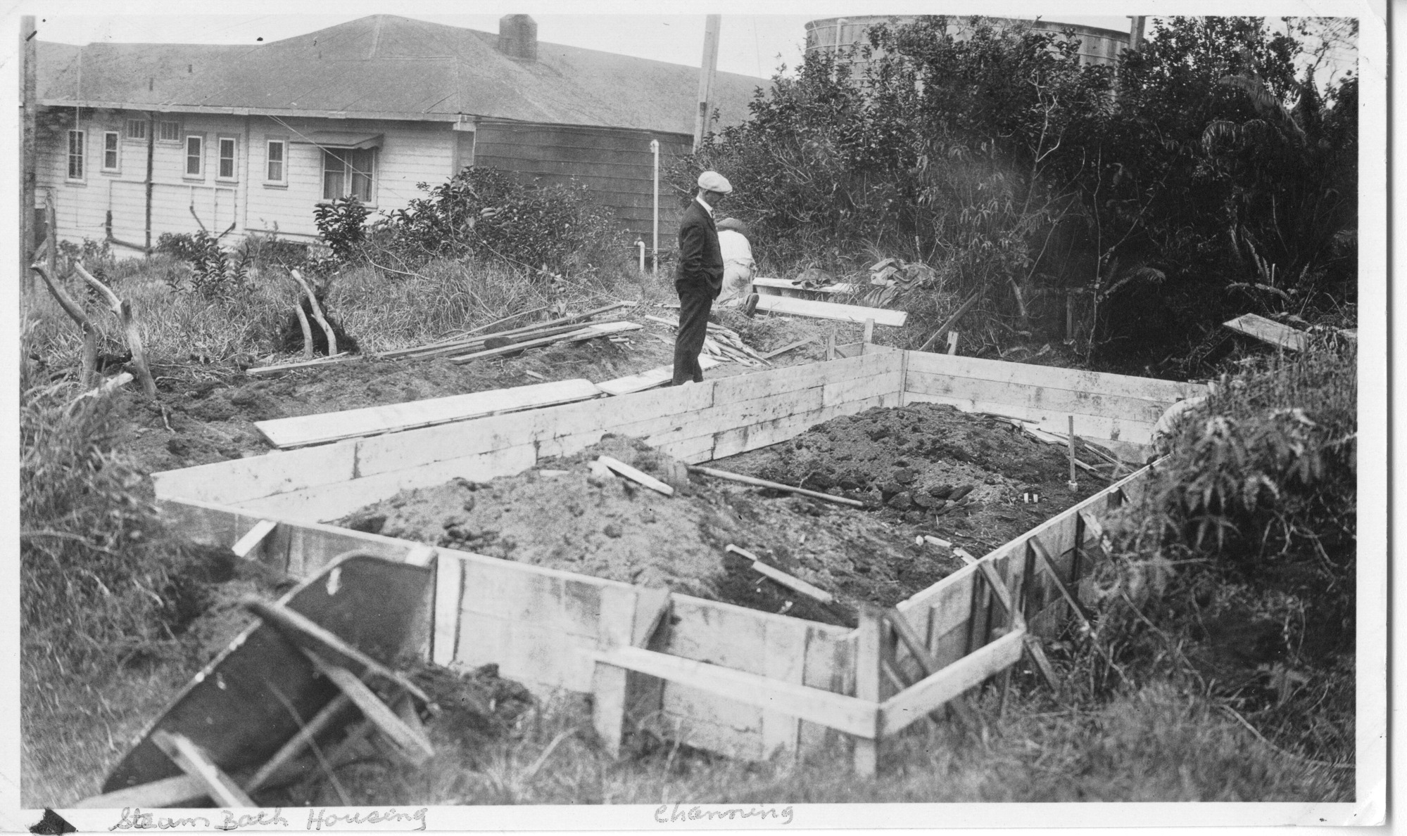 The image is in black and white. At the center of the image, wooden foundation propped up with supporting boards is filled with fine gravel and small rocks. A wheelbarrow sits tipped to its left side in front of the foundation. The area to the left of the foundation has been partly cleared, with tree stumps visible. There are some planks and boards on the ground there, with a worker near the back of the foundation kneeling on the ground, back turned to the camera. The rest of the construction site's surroundings are dense brush and dry grass. A man, Channing Lovejoy, stands behind the left edge of the foundation, hands in his pockets as he looks at the structure. He is wearing a suit and light-colored flat cap. A label written on the bottom white edge of the photo reads, "Steam Bath Housing, Channing."