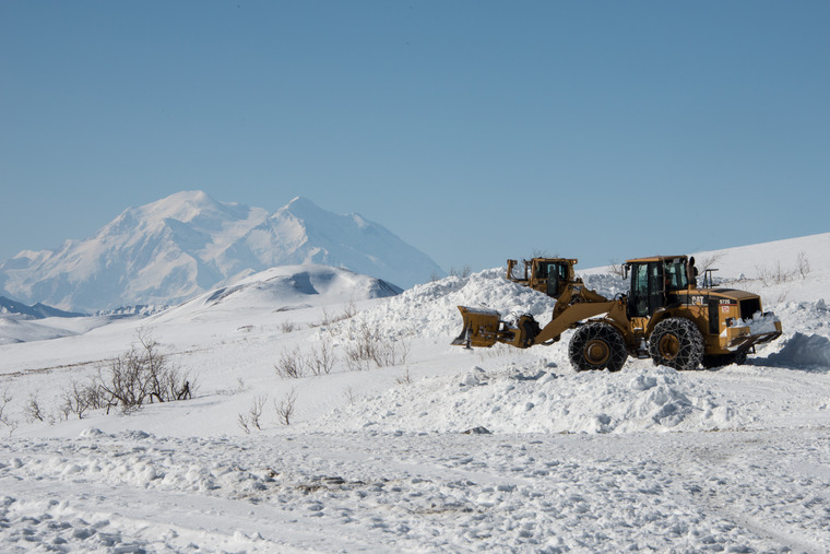 two front-end loaders plowing snow on a hill-top, overlooking a view of a huge mountain in the distance