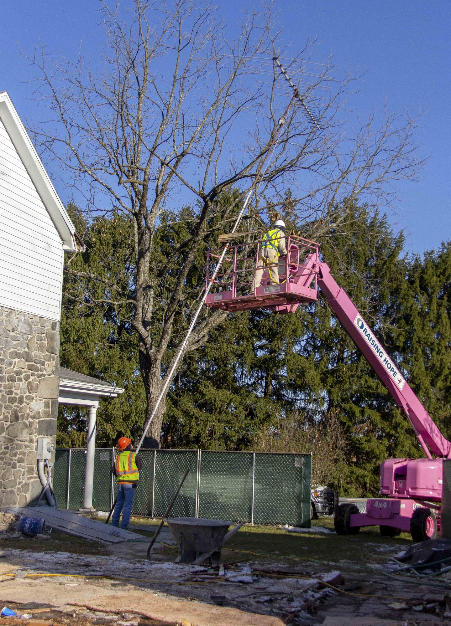 Two construction workers begin the process of removing the old TV antenna from the side of the house. One worker is holding the antenna at ground level and the other is navigating the atenna down through a tree while harnessed into the pink construction lift.