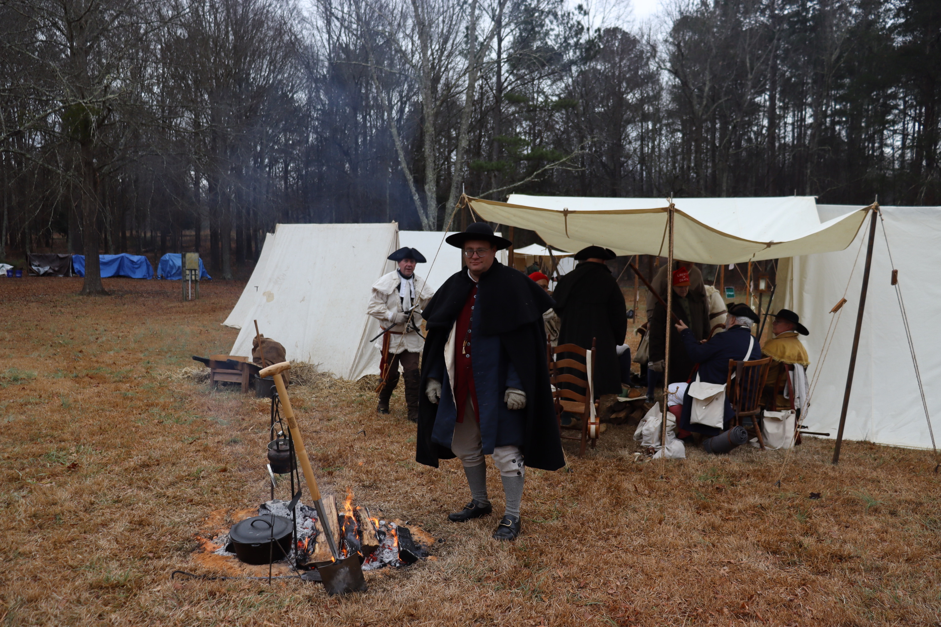A man stands near a fire with cooking materials over it. There are 3 tents behind him and some people talking.