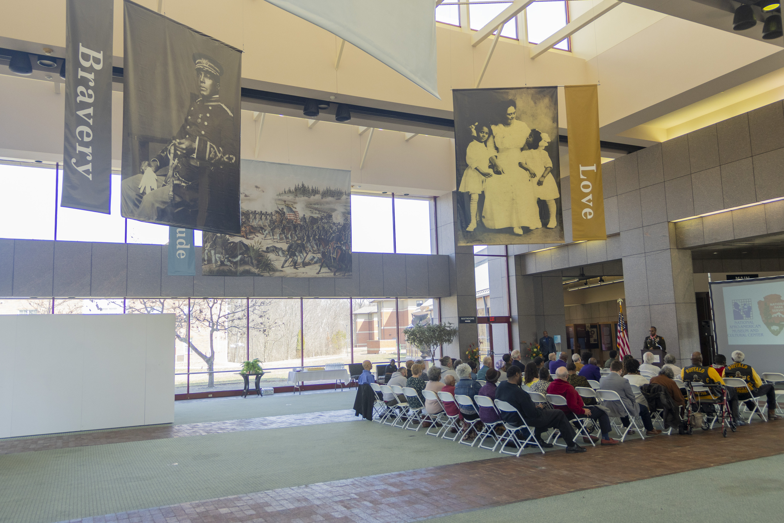 Seated guests in white chairs look and listen to a speaker who is on the far right, while a black and white picture banner of an African-American soldier hangs from the rafters above them all.