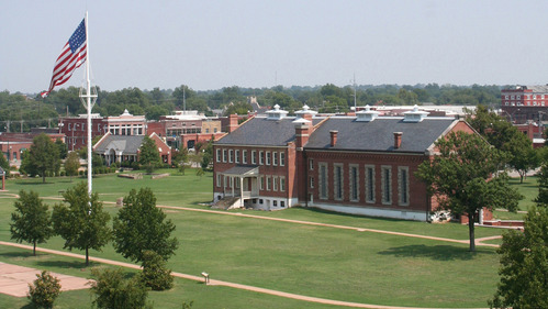Aerial view of a large 37 star flag flies on a 100 foot tall flag pole in front of the red brick visitor center on a clear sunny day.