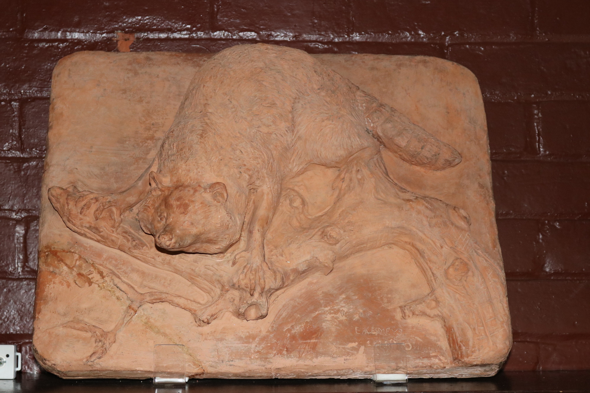A rectangular reddish clay plaque with a raised carving of a small mammal slinking on a tree branch. 