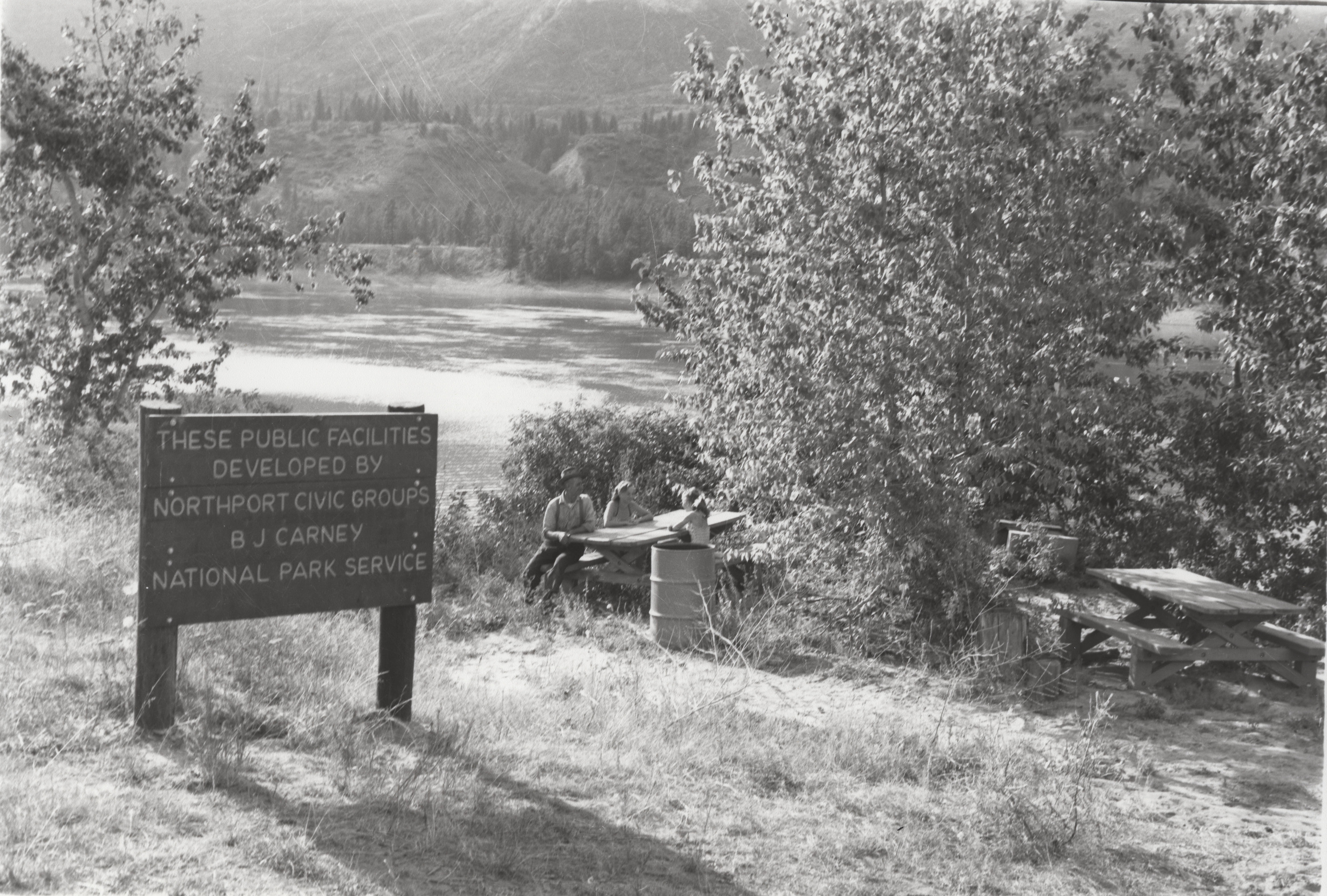Black and white photograph of a sign and picnic tables next to a lake