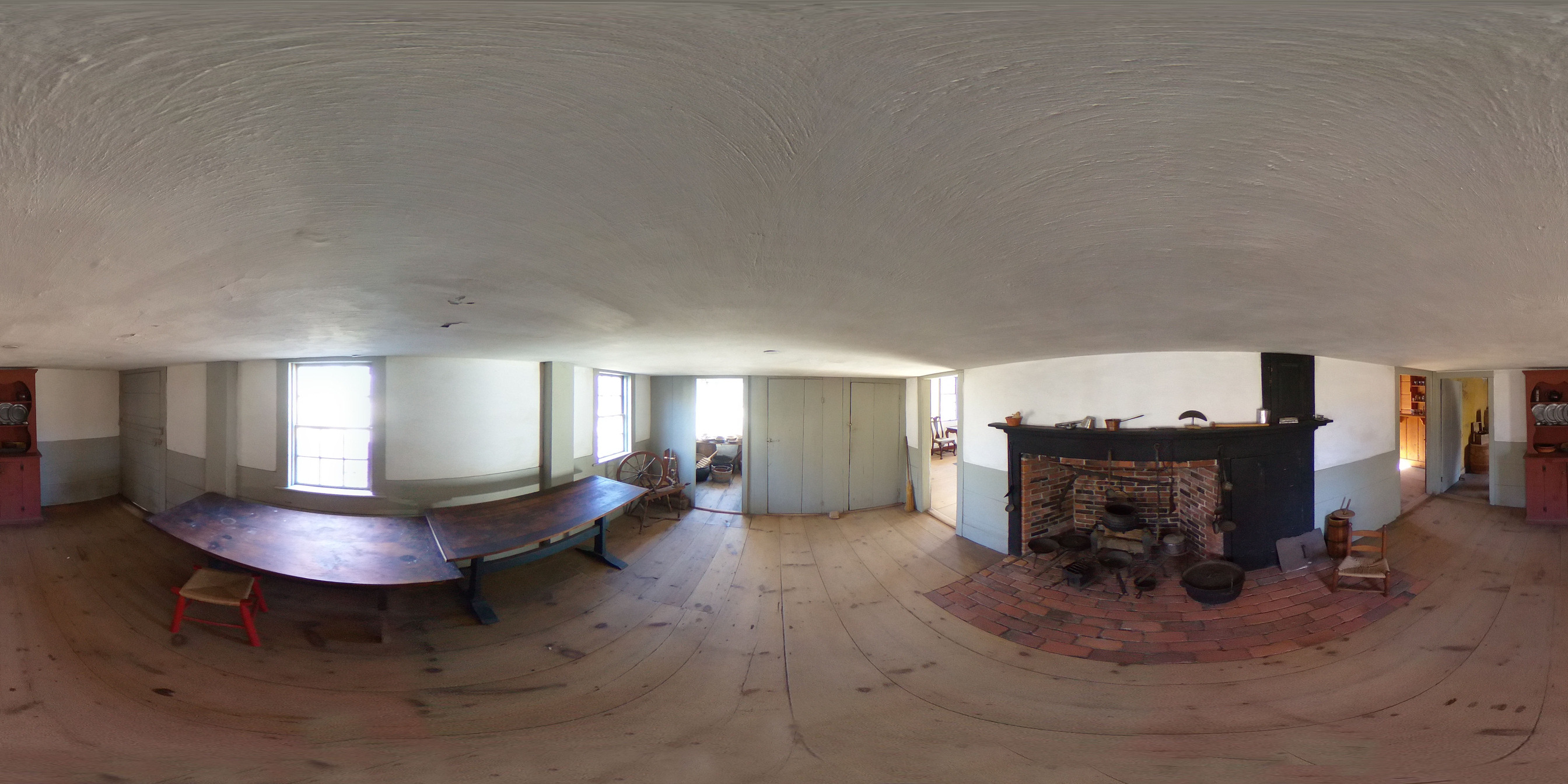 360 panoramic image of a long, narrow colonial room. The ceiling and walls are white with a sea green trim. The floor is unpainted. There is a large brick hearth set into the wall on the right with an oven on the side. Long tables line the back wall. Two small rooms are visible at either end. 