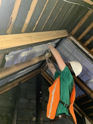 A worker in an orange safety vest installs white baffling panels among roof joists.
