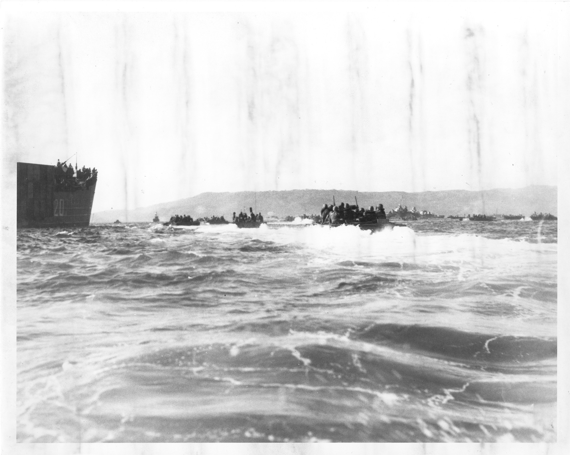 Small ships full of soldiers speed towards a beach. In the background are larger ships.