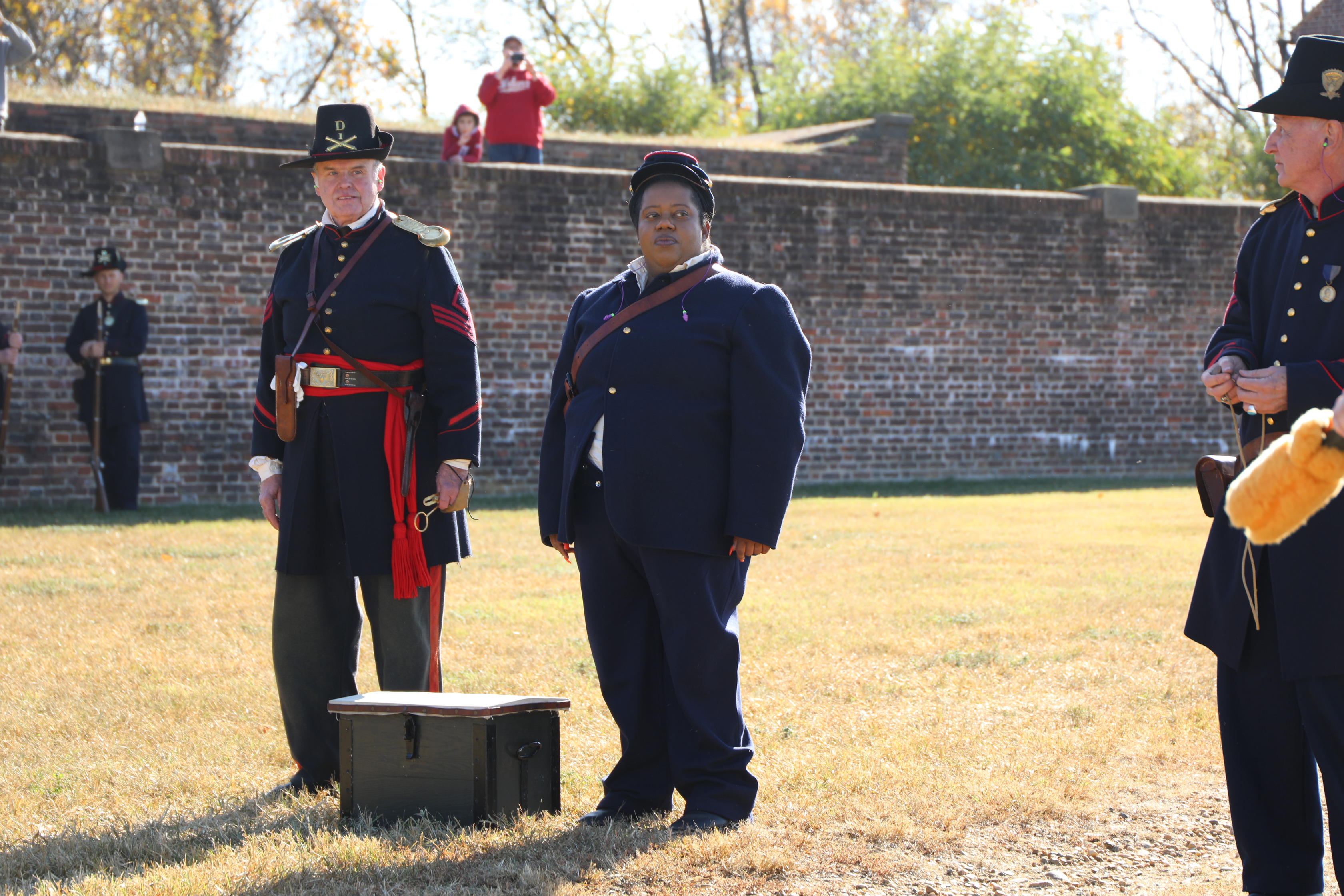 A person in a 19th-century military uniform, a dark blue coat, and a dark cap stands next to an open wooden chest. To their right, another man in a similar military uniform, but with a black hat, stands at attention. In the background, there's a brick wall, and behind it, a person in a red shirt can be seen. The setting is a historical artillery display on a sunny day.