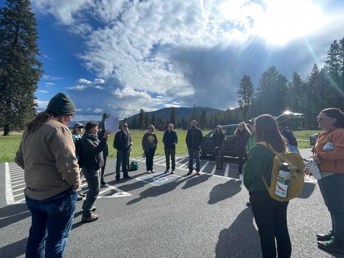 a woman stands, speaking to a group of people outside, in a parking lot, with conifer trees and green grass and blue sky in the background. 