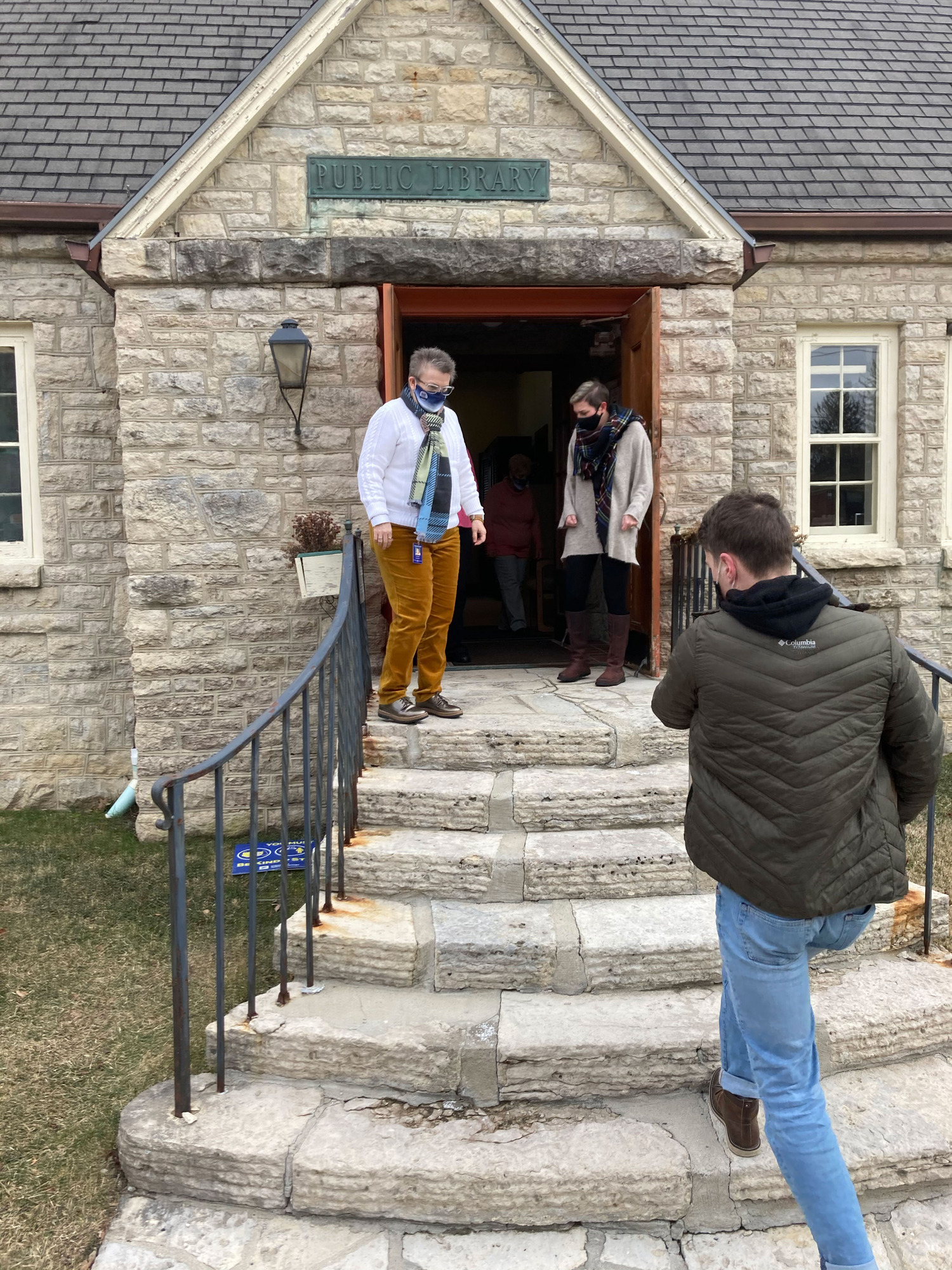 A man in a winter coat and holding a box walks up a small staircase while two women stand on either side of a doorway.