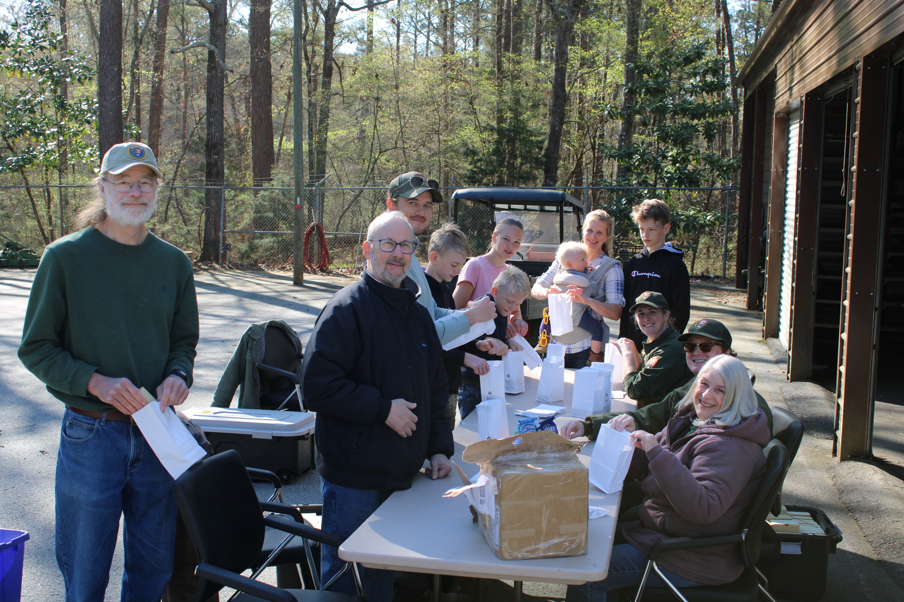 10 people stand and sit around a table filling white bags. 