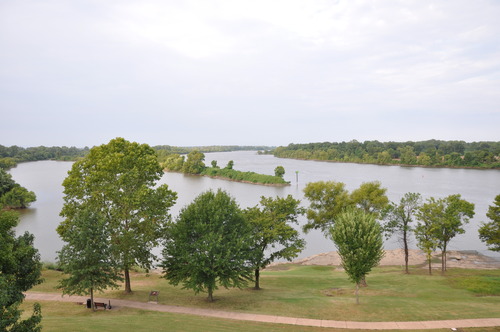 Two periwinkle rivers flow together, framed by bright green trees. In the foreground, the paved River Loop Trail winds through the grass.