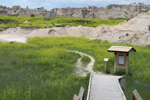 a boardwalk trail leads down stairs to a footpath meandering through prairie grasses and Badlands buttes in the distance