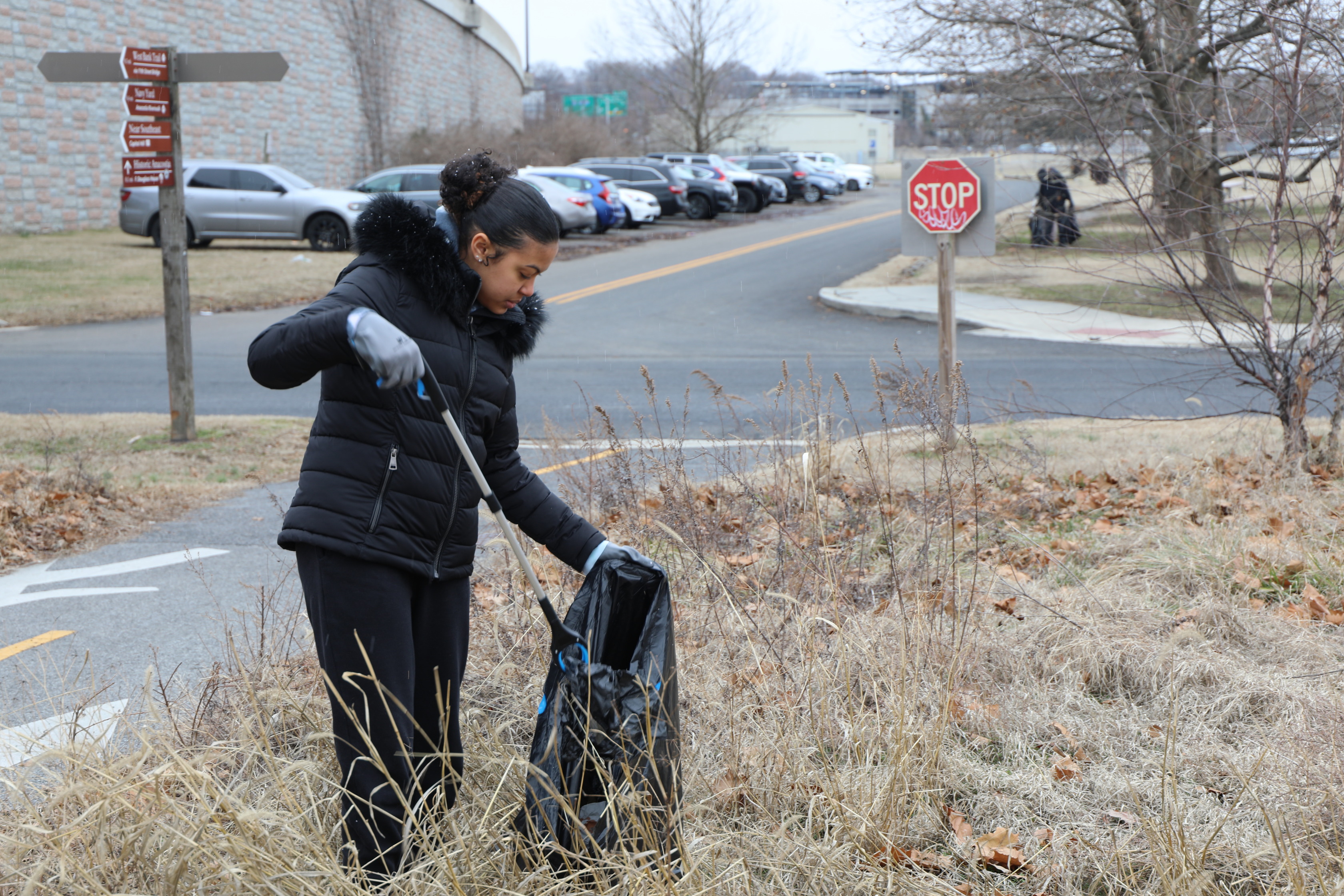 A young woman with her hair pulled back stands in a field of dry, tall grass and fallen leaves. She wears a black puffer coat with a fur-lined hood, black pants, and grey gloves. She is holding a long blue and white grabber tool in her right hand and is using it to put an item into a large black trash bag that she holds open with her left hand. To her left, there is a paved path and a wooden signpost with directional arrows. In the background, a parking lot with several cars is visible, next to a concrete wall. A red stop sign on a pole is also visible further down the road to her right. The sky is overcast, and the trees are bare.