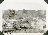 This is an historic black and white photograph from the Scotty's Castle Historic Photograph Collection, Death Valley National Park of a large l-shaped building with a flat roof setting along the length of a wash at base of barren desert mountains. Another building at right edge interrupted by plane prop. Number in black ink in lower left corner.