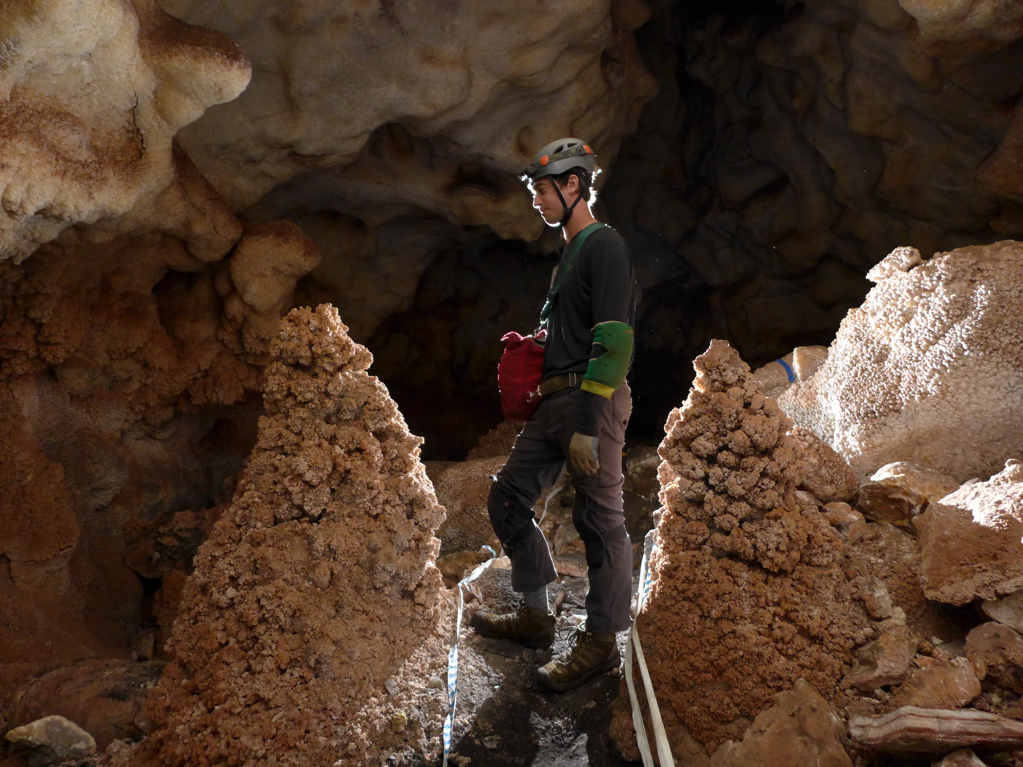 A man in a grey helmet and caving equipment stands in profile between two cave formations. 