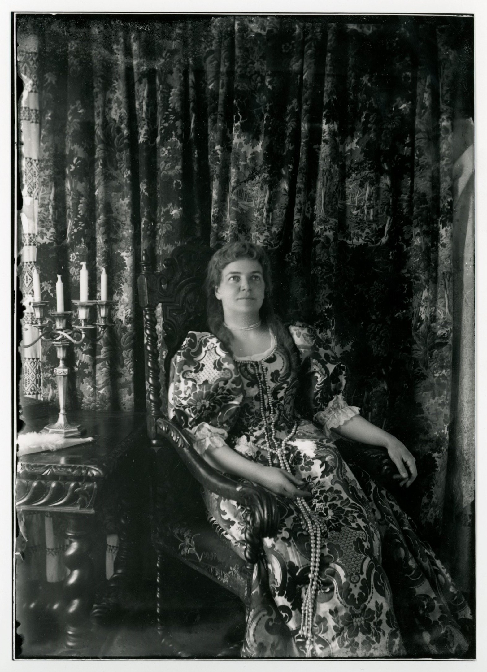 A white woman with long curls (perhaps a wig) sits in colonial-era gown next to table with candlestand. Very long string of beads falling below knees.
