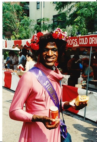 Marsha P. Johnson wearing a flower crown and Stonewall sash while holding a sandwich and soda in front of food vendors