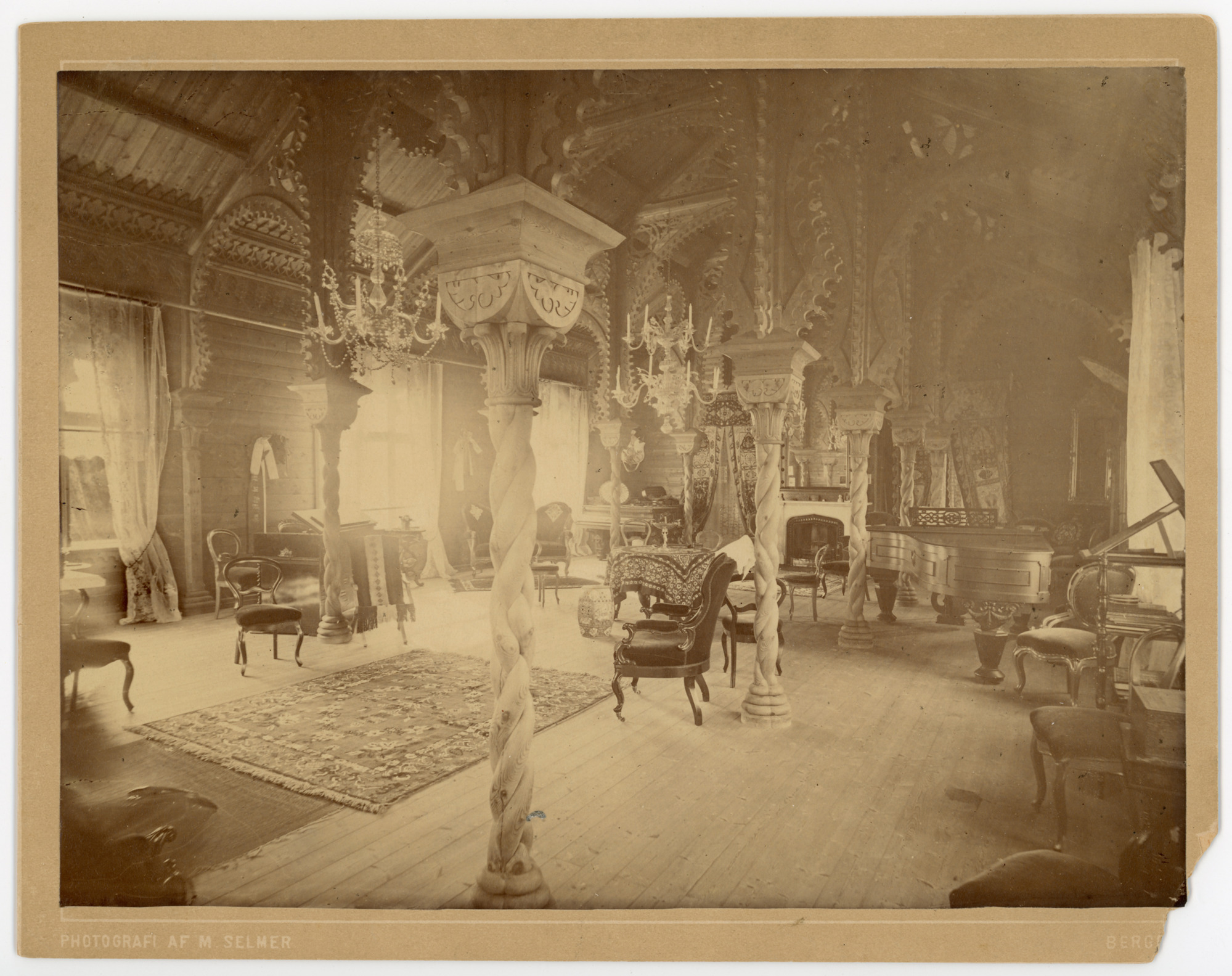 Interior view of room with piano to right. Twisted wooden columns supporting heavily carved arches line room. Two chandeliers, antique furniture, and carpet visible.