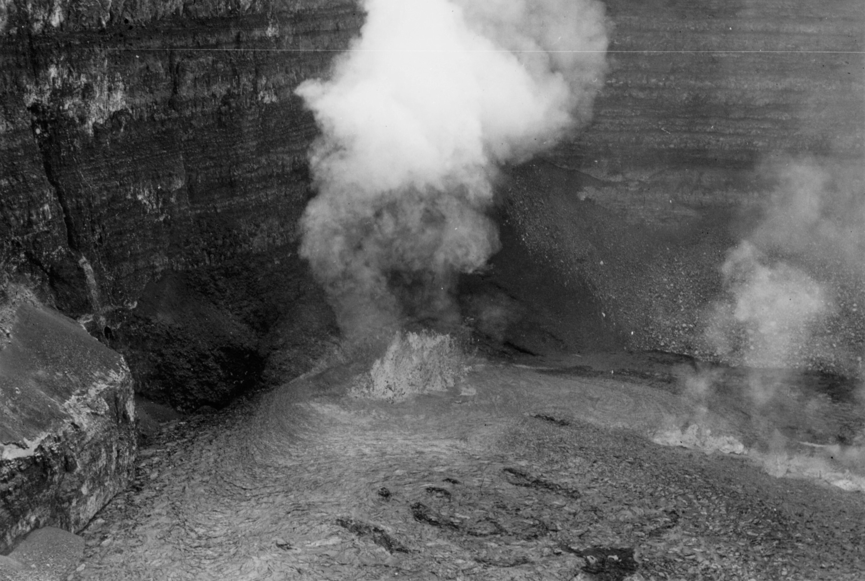 Black and white image of a large lava fountain emitting steam along the bottom edge of an erupting crater. 