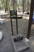A plastic panel with information on it sits inbetween metal legs. Behind it are trees, logs, and rocks. 