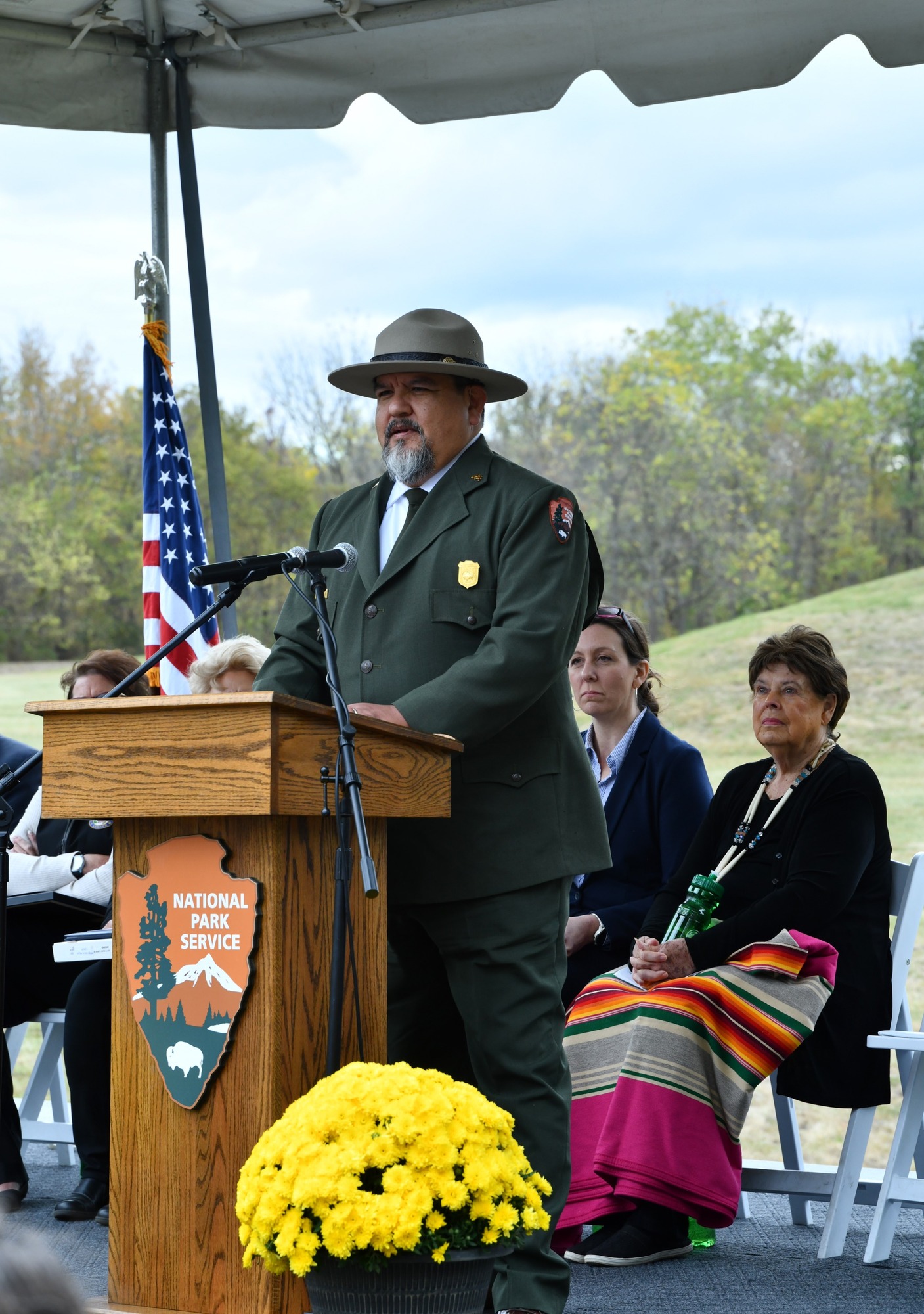 A man wearing a green suit with green tie and tan, flat-brimmed ranger hat stands behind a wooden podium and speaks in to a microphone.