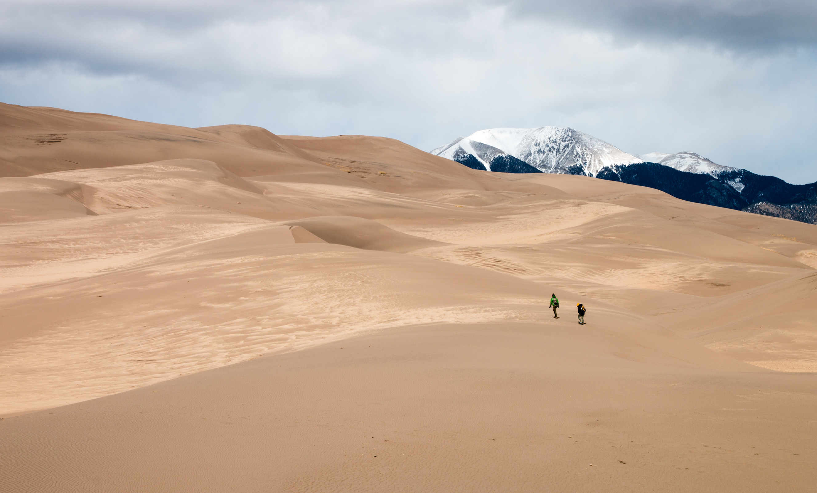 A couple of park visitors walk up the dunes. In the distance the snow-capped Sangre de Cristo Mountains rise above the dunes. 