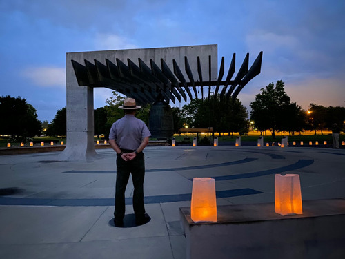 A person in uniform stands in front of a large bell suspended from a concrete structure. Luminarias are aglow around the bell. 