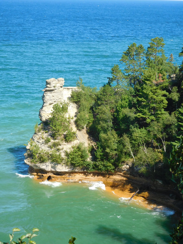 Eroded sandstone rock formation. The top harder rock looks like a castle turret. Trees line the top and sloping sides of the cliff and the Castle sits at the end with Lake Superior at its base.