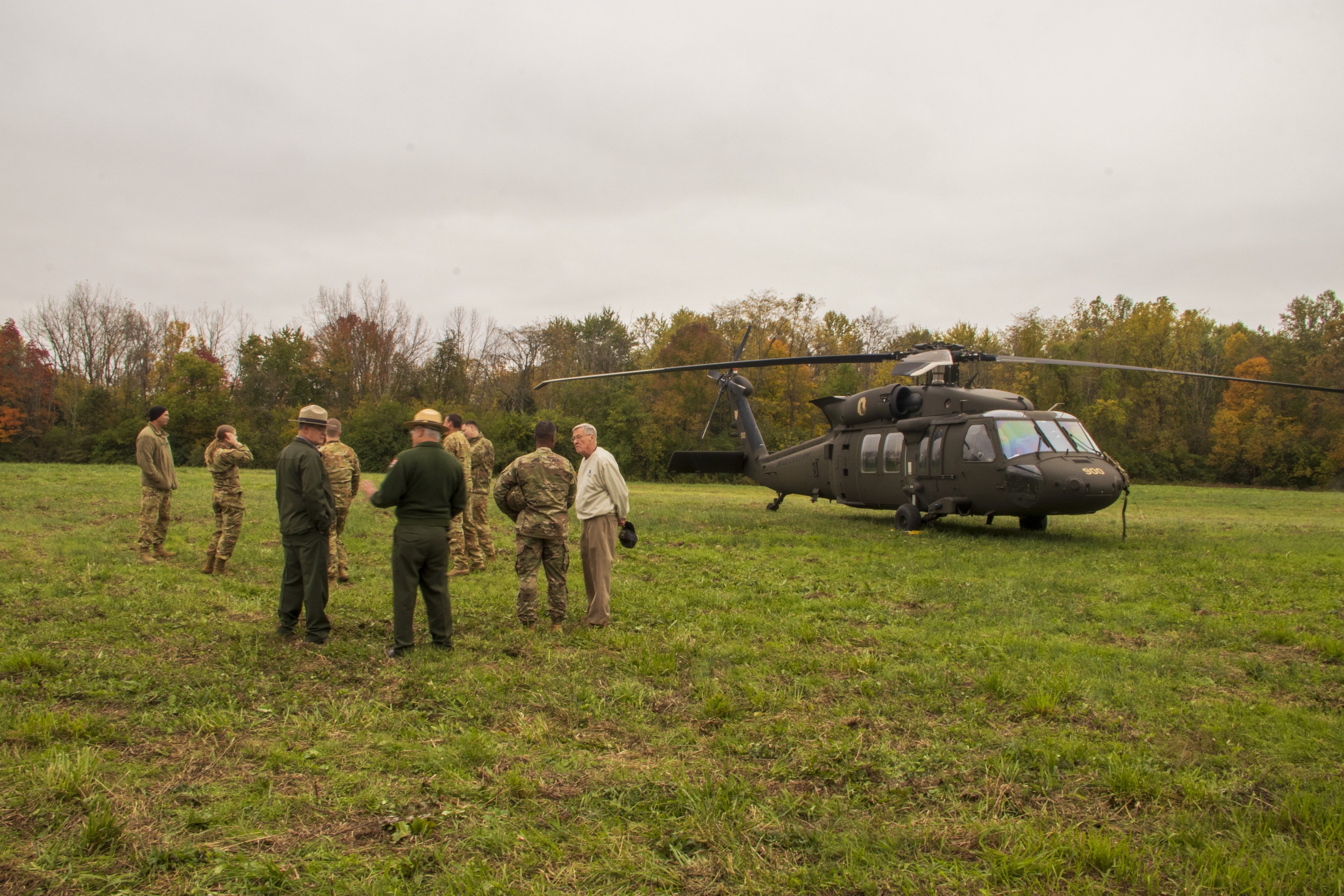 Several people in tan-colored camouflage uniforms and two park rangers with flat hats stand to the left of a parked helicopter in a grassy field.