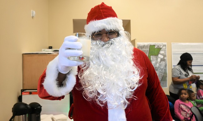 A Black man dressed as Santa wears a silk red hat trimmed with white fur, a red velvet jacket also trimmed with white fur, white glove, round glass, and a long white beard. He holds up a cup of milk in a clear glass towards the camera, a twinkle in his eye. Children behind him make decorations at their table while one looks at Santa. 