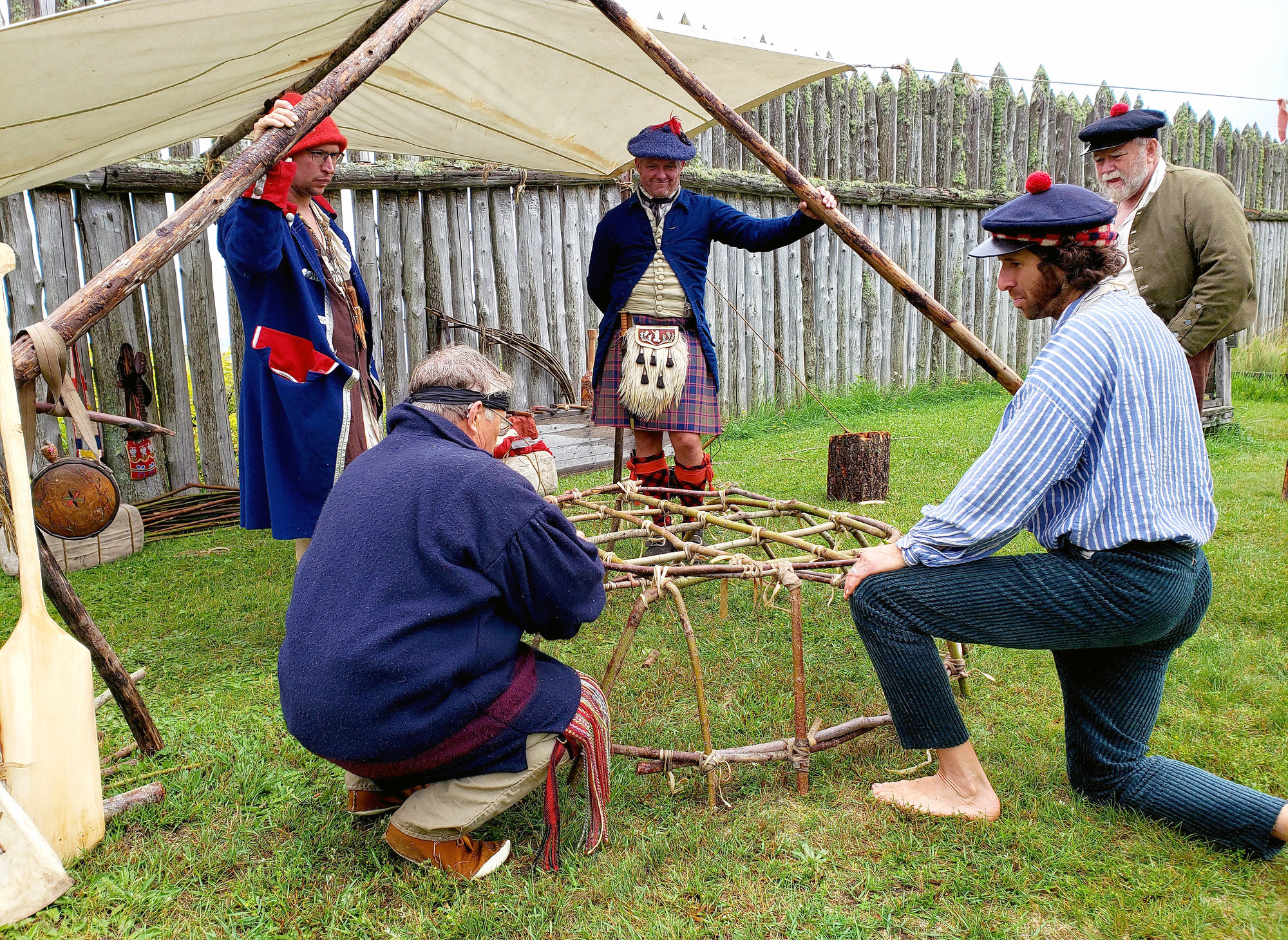 A group of five people wearing historic clothing. Two are tying crossed willow branches together to make a bowl shape.
