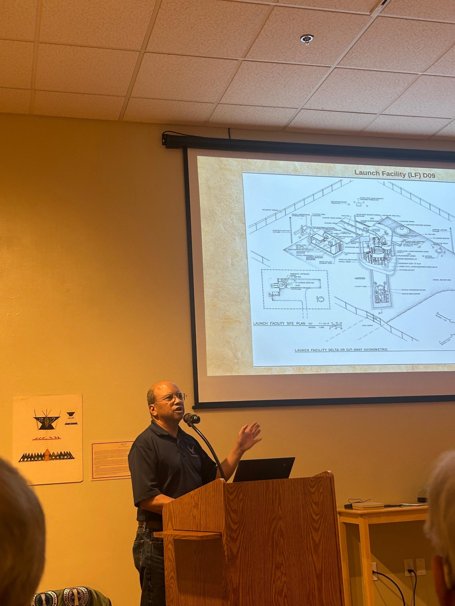 Man in a blue shirt at a podium speaking into a microphone. A large black and white diagram is projected onto the screen beside him.