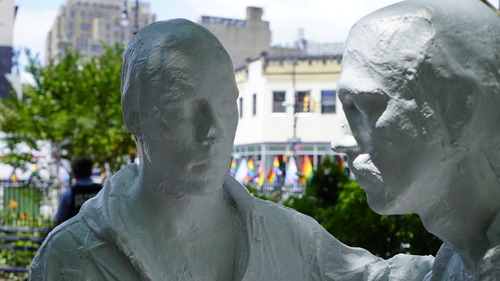 Two white plaster and stone sculptures depicting the heads and necks of human figures. In the background are blurry green trees and rainbow flags 