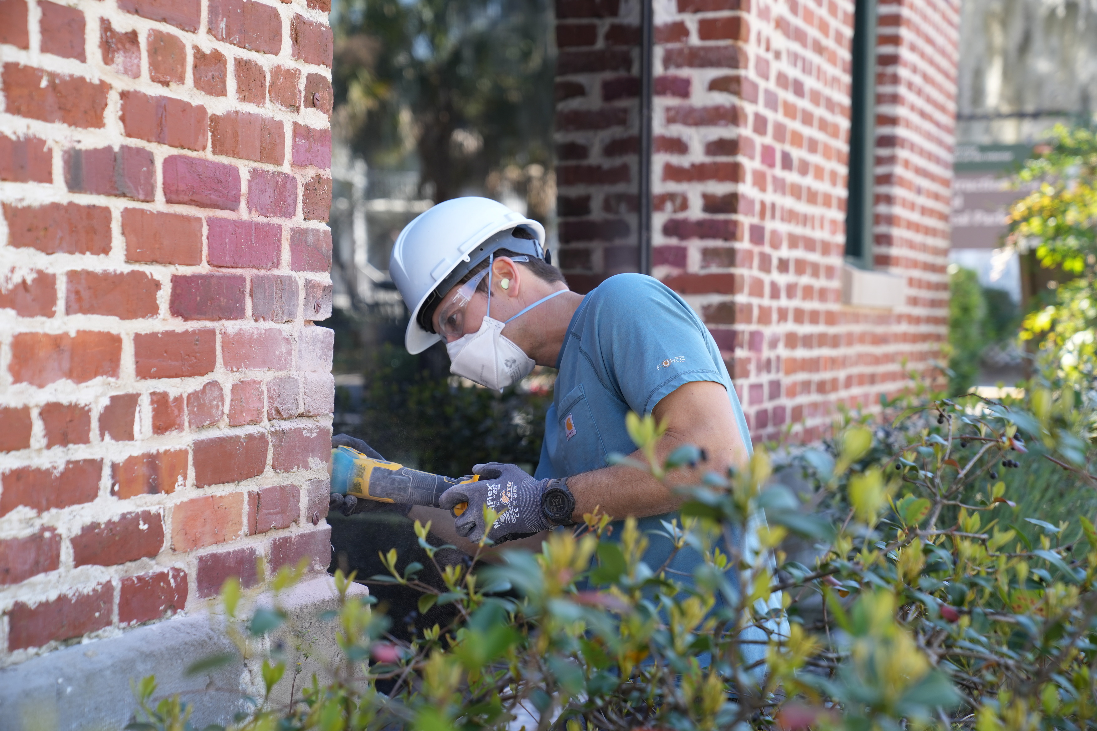 A man wearing personal protective equipment uses a power tool to drill out mortar between bricks.