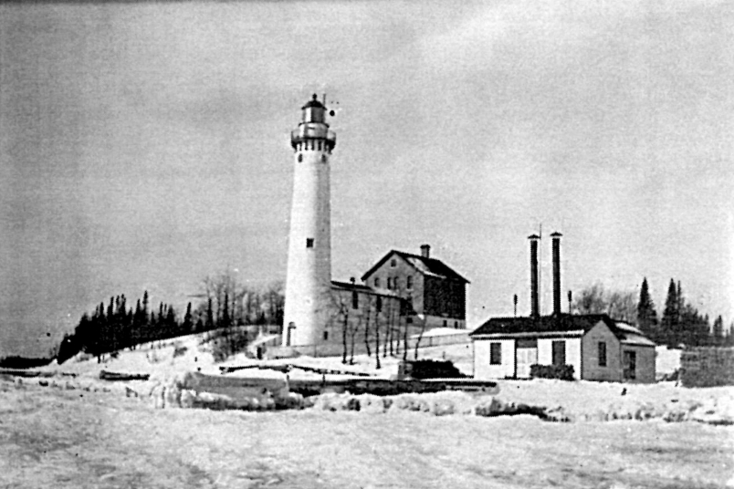 A black and white image of a tall cylindrical building on a small hill with a small building in the shape of a T to the left.