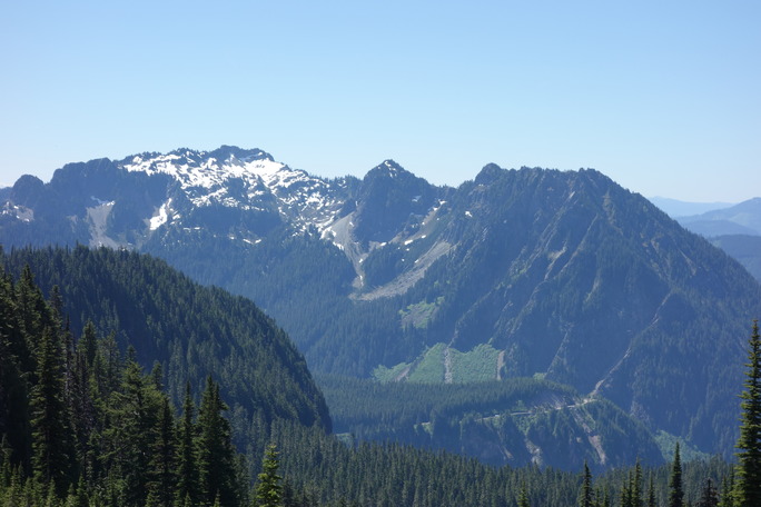 A mountain peak at the end of a range of peaks, covered in forest with patches of subalpine meadows on the lower slopes and snow around the summit. 
