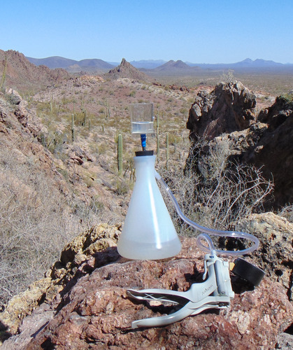 A plastic bottle with a cylinder attached to the top sits on a rock above a desert landscape with saguaros and mountains. Also attached to the bottle is flexible tubing attached to a metal instrument designed to be squeezed by a hand.