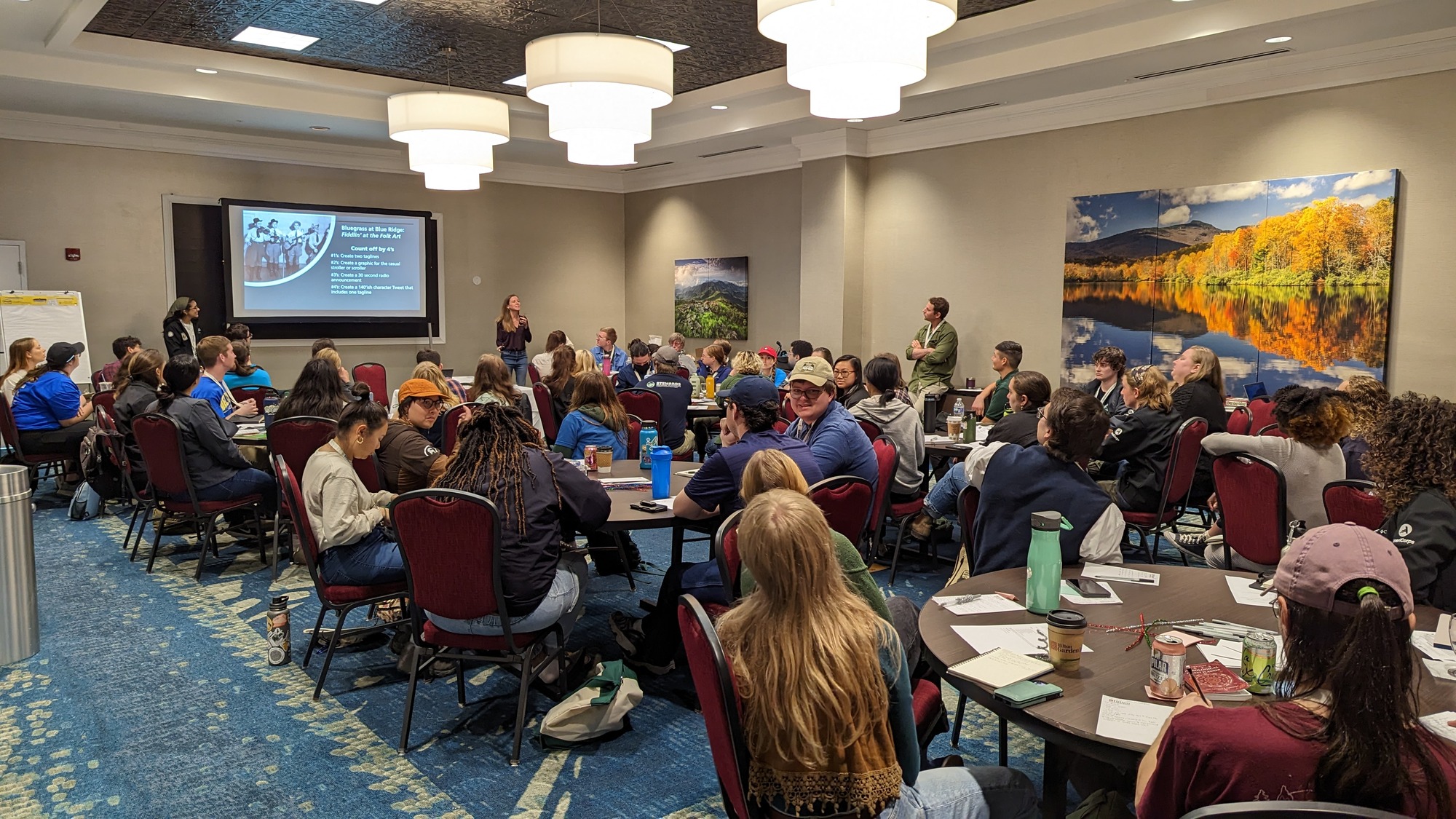 a large group sitting at round tables in a meeting hall, a speaker stands at the front of the room.