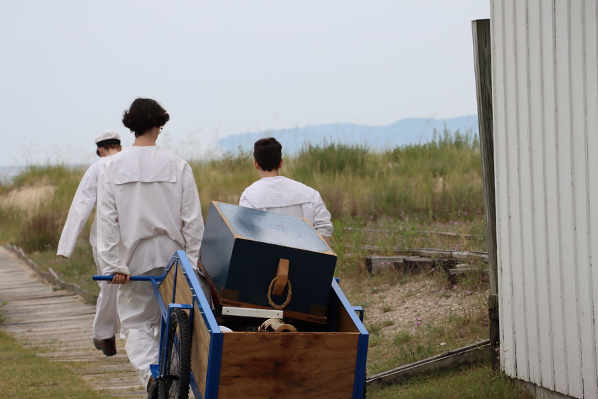 Surfmen Rachel, Merrick, and Britt, clad in their dashing white uniforms, haul the beach apparatus loaded with lifesaving equipment down to the beach to perform a daring rescue.