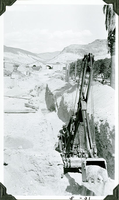 This is an historic black and white photograph from the Scotty's Castle Historic Photograph Collection, Death Valley National Park of laborer operating power shovel to excavate tunnel in front of Scotty's Castle Hacienda. Tunnel known as Multipurpose Facility (MPF) Tunnel. Looking east. May 1931. Photographed by Mat Roy Thompson.