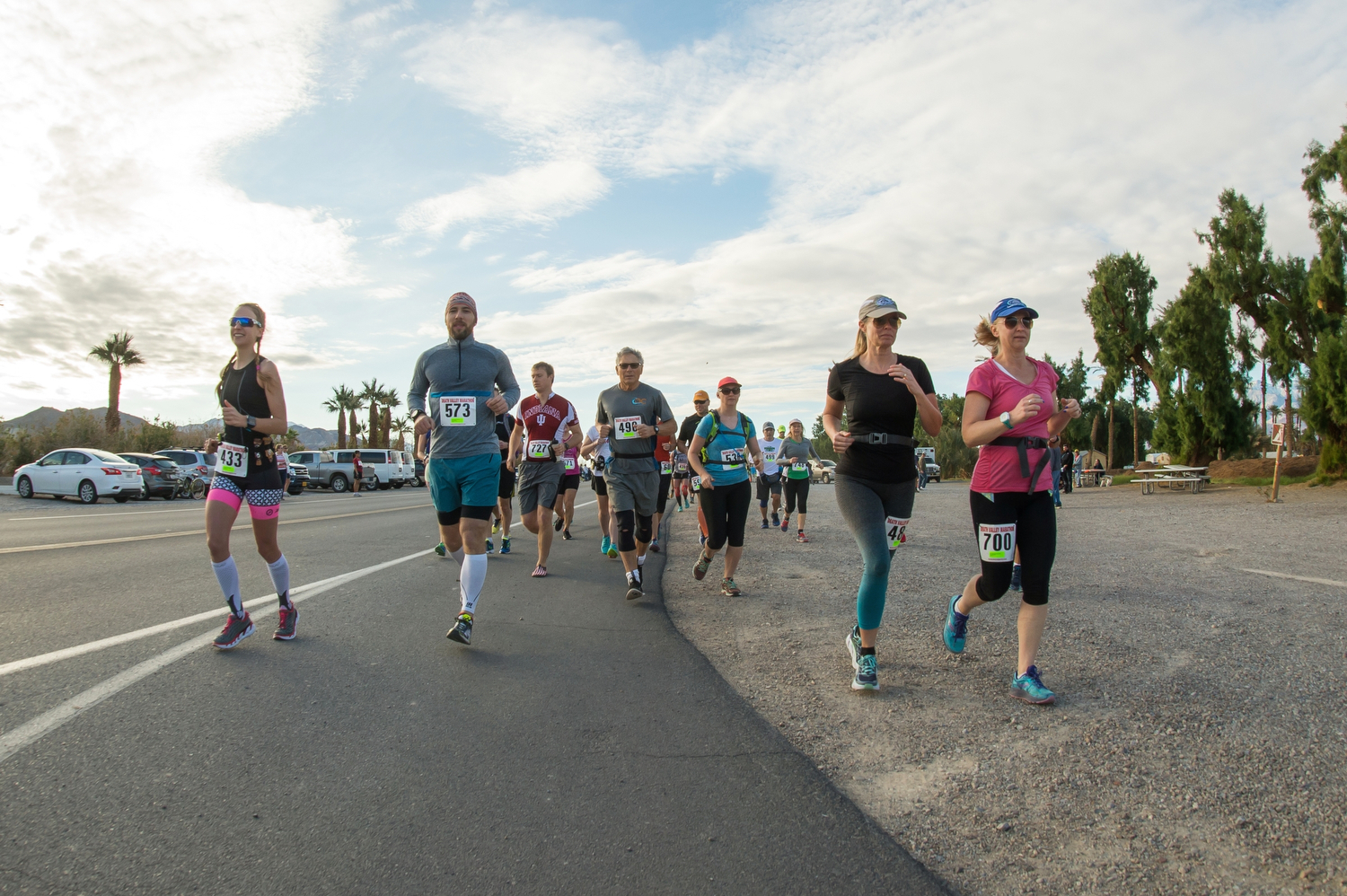 A large group of runners with numbered stickers run towards the photographer on the side of the road, with palm trees and a partly cloudy sky.