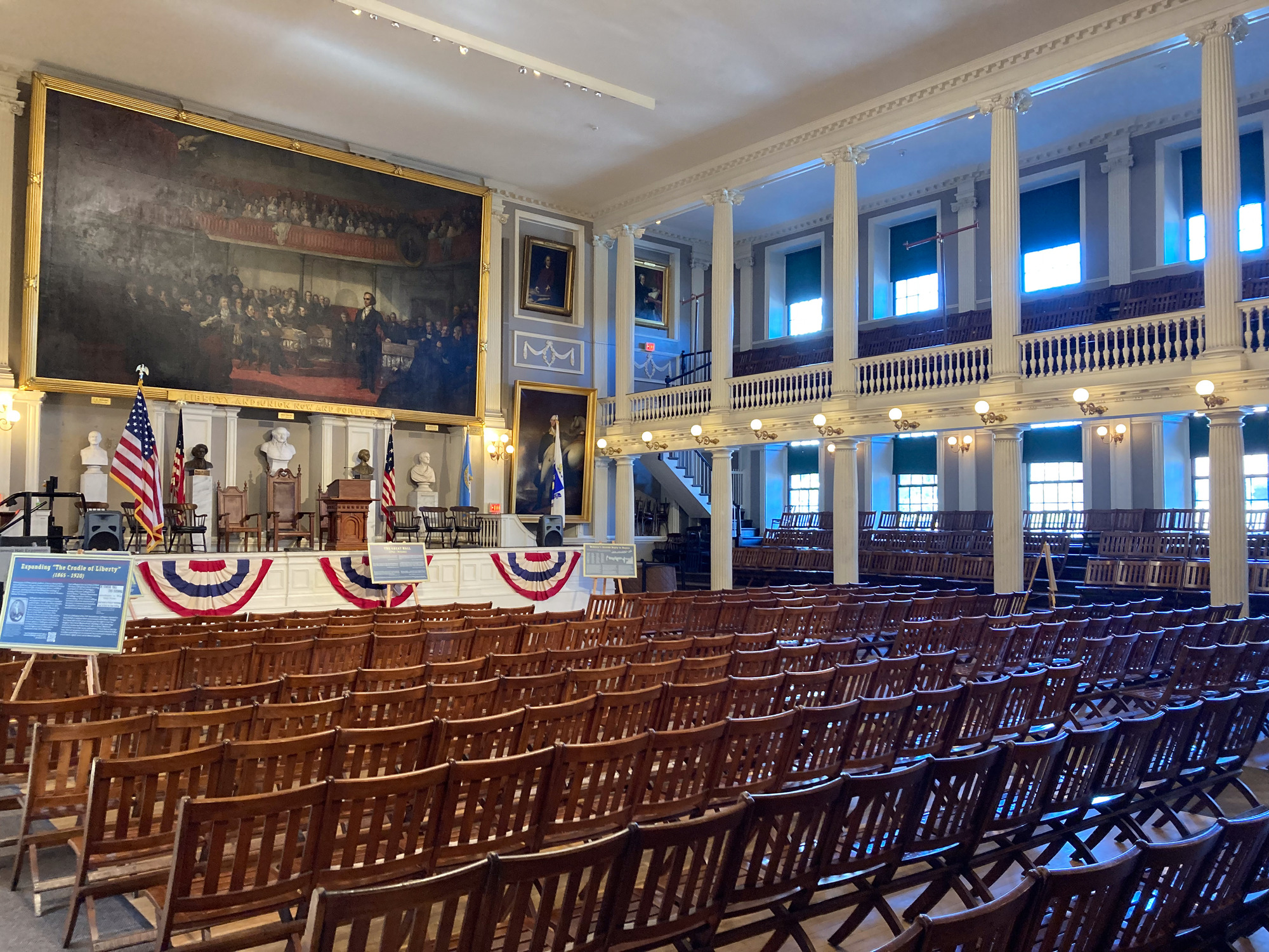 View of the interior of the Great Hall, in the foreground are brown wooden chairs that are attached to each other facing a white stage with bunting across the front and a massive painting of the old senate meeting chamber above it. Above the chairs the white balcony with more chairs looks down upon the stage.