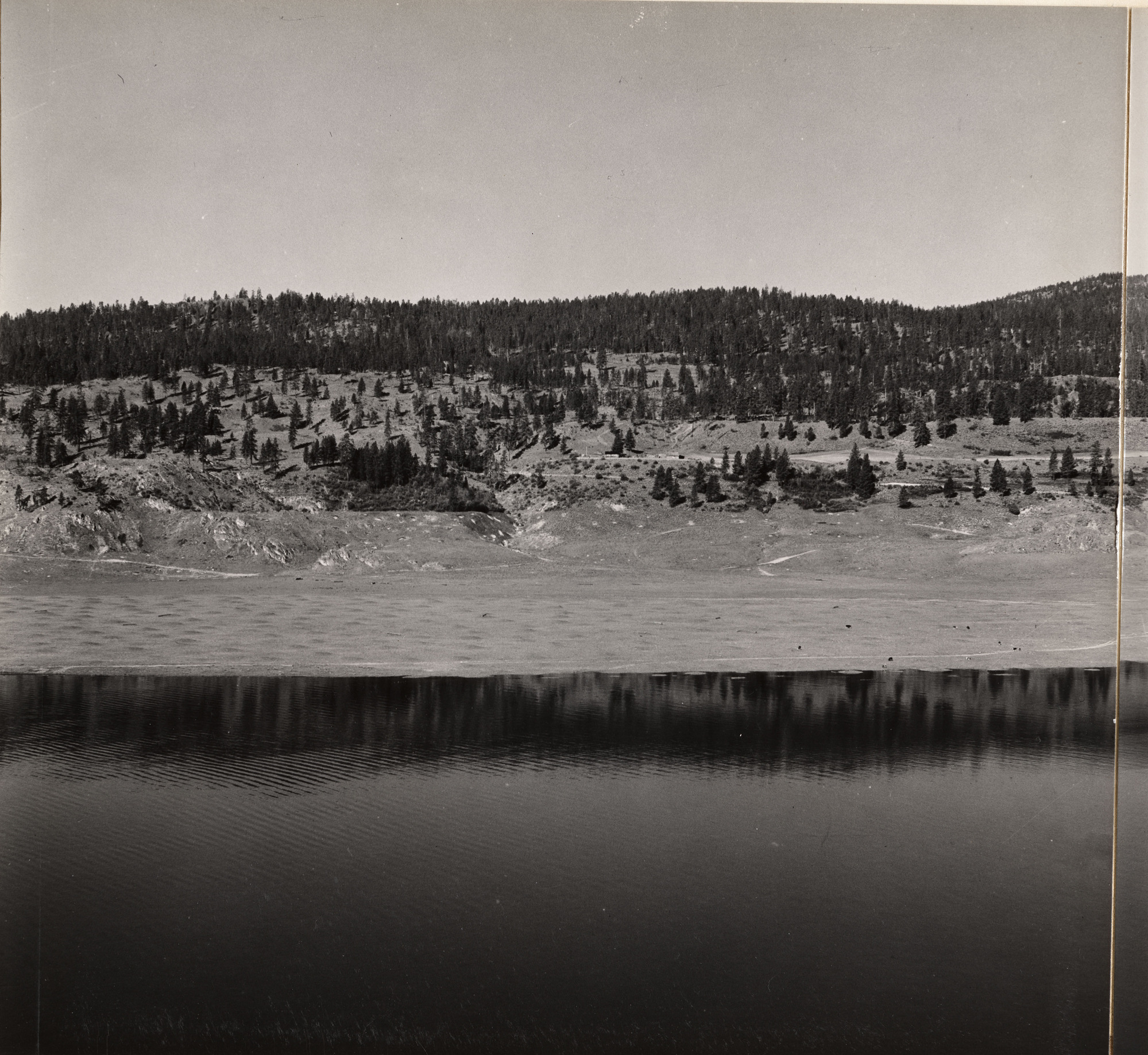 Black and white photograph of the hilly forested shore of a body of water.